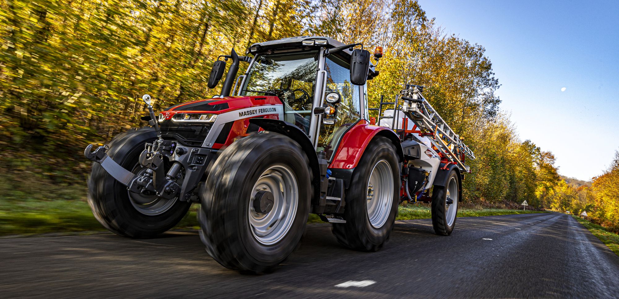 Ein Traktor fährt eine Straße entlang, im Hintergrund sind Bäume zu sehen.